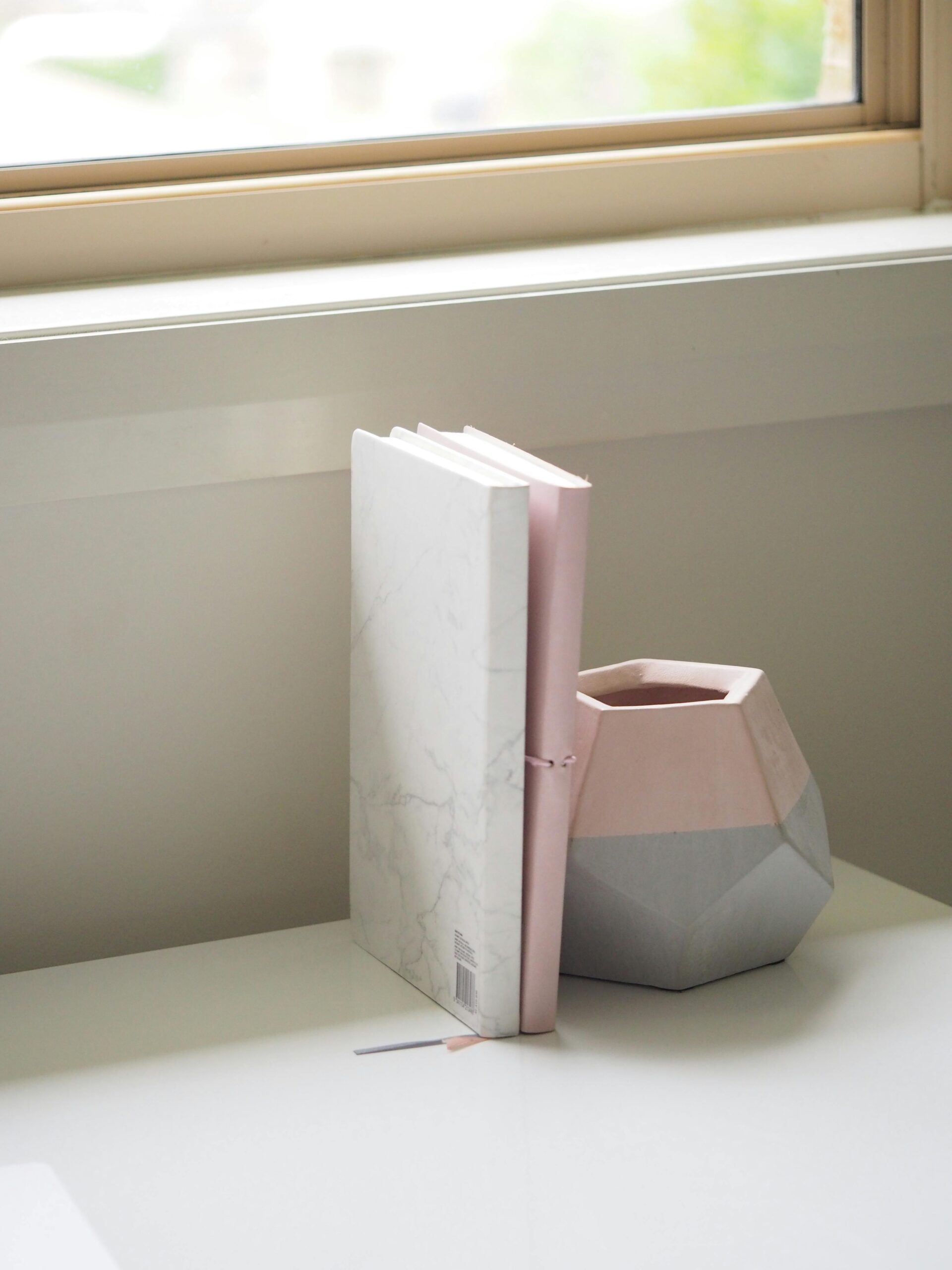 Simple and elegant desk scene featuring a marble notebook and a geometric vase by a window.