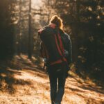 Full body back view of anonymous female hiker with rucksack strolling in woods with tall trees in nature during trekking